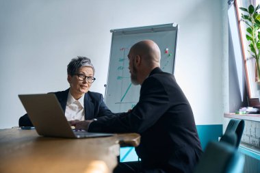İki orta yaşlı profesyonel, bir ofisteki ahşap bir masada iş birliği yapıyor. Gri saçlı ve gözlüklü kadın dizüstü bilgisayarına odaklanırken konuşuyor ve adam beyaz tahtadaki bir grafiği inceliyor..
