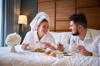 Genç çift yatakta keyifli bir kahvaltı yapıyor, gülümsüyor ve lezzetli yemekler yiyor. Romantik atmosferi geliştirmek için bir vazo çiçek içerir..