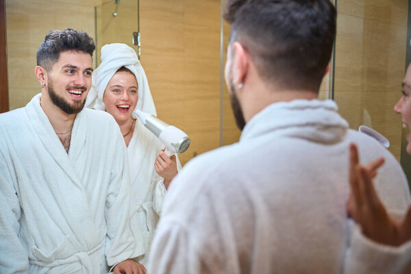 Three young adults, two men and one woman, enjoy a playful moment in a bathroom. The woman with red hair aims a hair dryer at one man while they all laugh in white bathrobes.