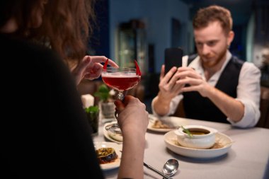 Yetişkin bir çift modern bir restoranda samimi bir anı paylaşıyor. Kadın elinde bir kokteyl tutarken düşünceli görünüyor ve erkek de yemek pişirme deneyimini telefonuyla yakalıyor..