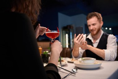 Yetişkin kadın elinde kırmızı garnitürle kokteyl tutarken gülümseyen adam akıllı telefonuyla fotoğraf çekiyor. Sahne, bir yemek ortamında neşeli ve rahat bir atmosfer yakalar..