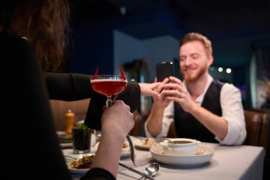 İki kişi bir restoranda keyifli bir akşamı kutluyor. Biri elinde şeytan boynuzlarıyla kokteyl tutarken diğeri gülümsüyor ve o anı akıllı bir telefonla yakalıyor..