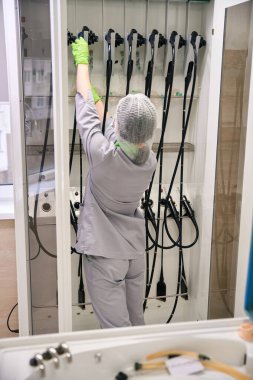 Eldiven ve saç filesi de dahil olmak üzere tıbbi kıyafet giymiş beyaz bir kadın steril bir depoda tıbbi malzemelerle etkileşime giriyor. Gündüz odaklı ve düzenli atmosfer.