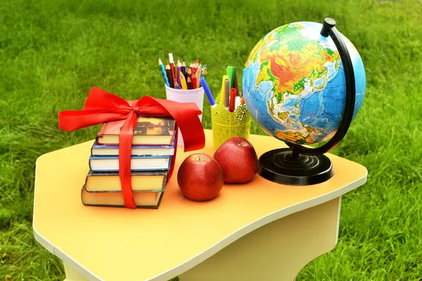 On a small school table against a background of green grass - books tied with a red gift ribbon next to a globe, multi-colored pencils and two red apples