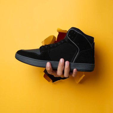 Sağ insan eli, siyah spor ayakkabı tutarak sarı kağıt duvarı yardı. Erkek eli spor ayakkabısı tutuyor. Spor ayakkabıları tutan ellerin modeli. Reklam ihtiyaçlarınız için uygun. Güzel ayakkabılar.                               