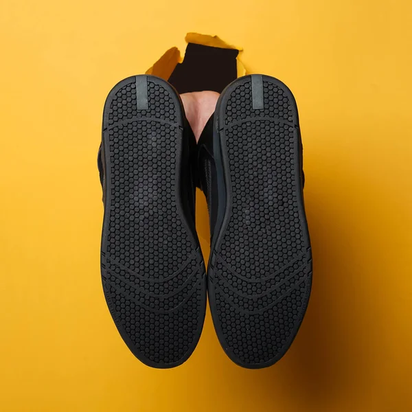 the man's right hand holding a pair of shoes through the yellow paper background. Copy space for ad content. Best choice concept. Holes in wall paper. Mock Up of creative advertising space. Sneakers.