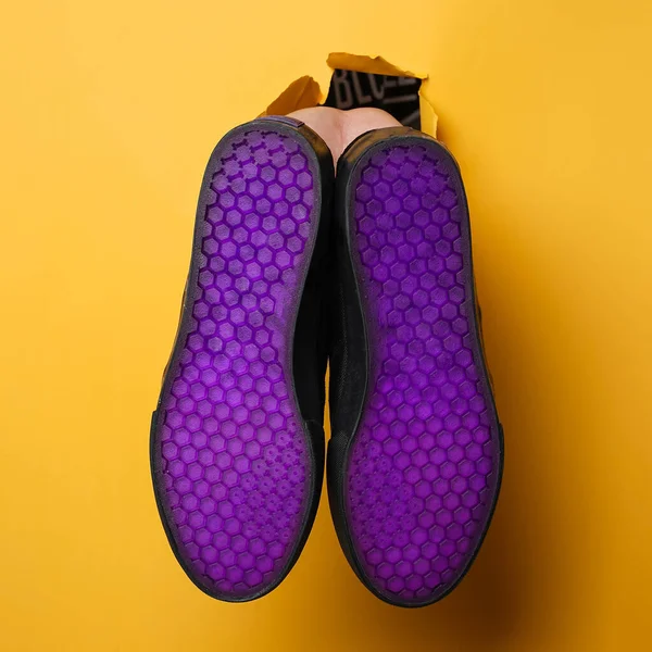 Plain black sneakers with a purple base. Plimsoll shoes on a yellow background give a bright effect to the photo. Front shot. Side shot. Hands holding sneakers through yellow paper background.