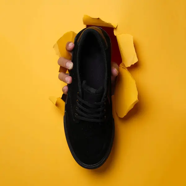 Plain black sneakers with a purple base. Plimsoll shoes on a yellow background give a bright effect to the photo. Front shot. Side shot. Hands holding sneakers through yellow paper background.