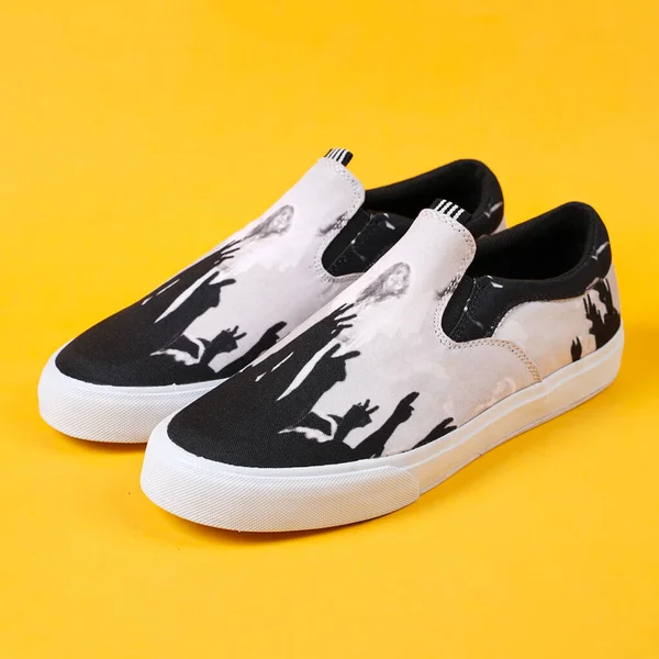 Pair of black shaded white sneakers with white edges all over, facing the sides. The color combination of white slip-ons against a yellow background creates a gorgeous color contrast. Shooting the camera from the front makes the shoes look beautiful.