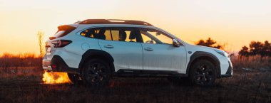 Brno, Çek Cumhuriyeti - 10 Şubat 2026: Beyaz Subaru Outback SUV, kışın gün batımında bir arazide. Akşamları gökyüzü turuncu ışıkla aydınlatılıyor. Japon bir jiple..