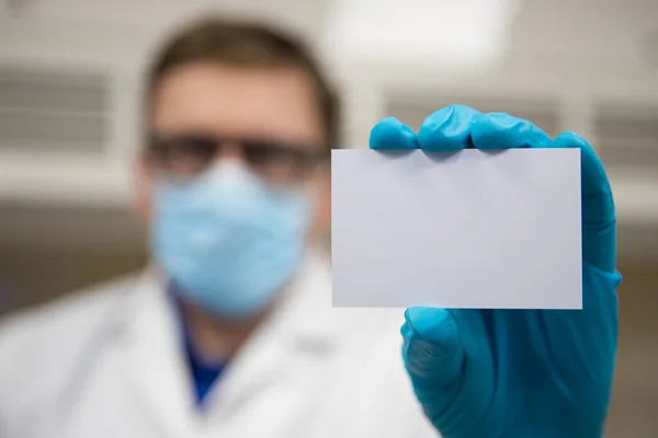 Close up of doctor holding a blank business card. Scientist wearing a lab coat, face mask and protective gloves