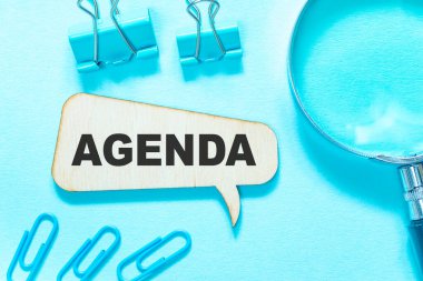 Gündem, kelime ahşap bir kutunun üzerine yazılmış mavi arka planda, kırtasiye malzemesi etrafa yayılmış..