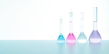 İçindeki renkli sıvılarla aynı hizada duran açık cam şişeler, kimyasal maddeler, çözümler, laboratuvardaki beyaz masanın üzerinde duruyor..