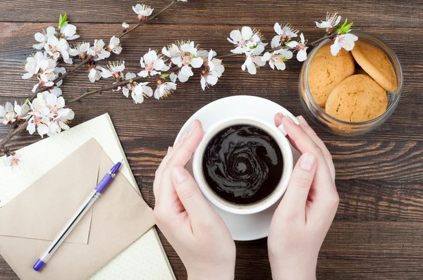 Mani donna che tengono la tazza di caffè su sfondo di legno primavera. Lettera, carta bianca, penna, fiori, tazzina da caffè e biscotti sul tavolo di legno — Foto Stock