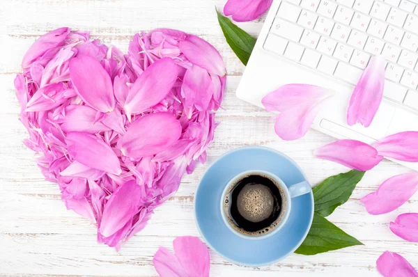 Spazio di lavoro con computer portatile, tazza di caffè e fiori su sfondo in legno. Petali di peonie a forma di cuore — Foto Stock