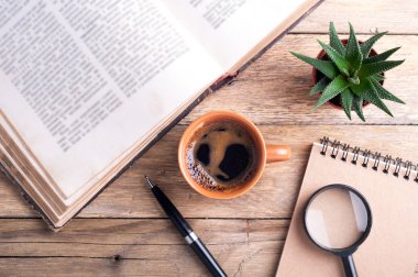 Bir fincan kahve, eski açık kitap, etli, kalem, defter ve Büyüteç rustik ahşap zemin üzerinde. Ofis Resepsiyon Masası