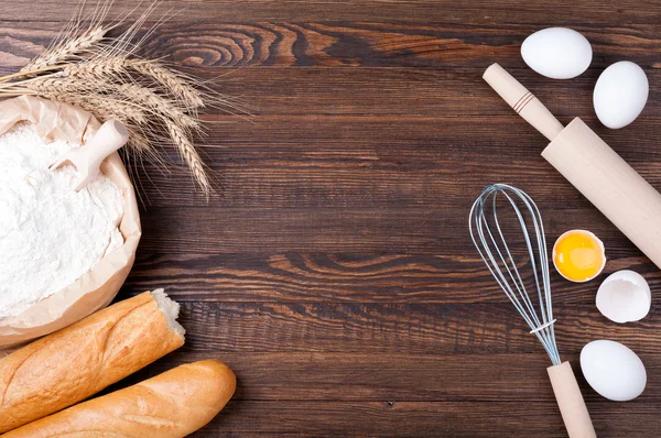 Flour in paper bag, wheat and baguette on wooden background. Ingredients for baking - flour, eggs and kitchen utensils rolling pin and whisk