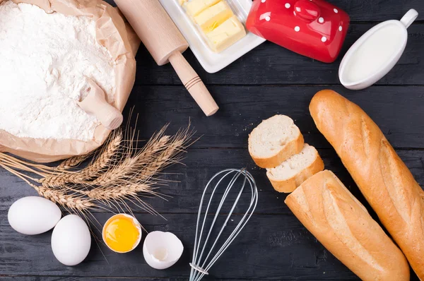 Baking background. Flour in paper bag, eggs, butter, milk, rolling pin and whisk on dark wooden background. Ingredients for cooking homemade bread — Stock Photo, Image