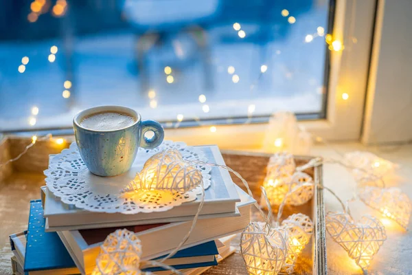 Coffee cup, books, sparkle fairy lights on window sill on blurred background. Christmas season. Concept warm and cozy home, reading books, winter holidays, hygge, evening rest and comfort