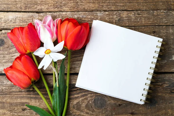 Bouquet of tulips and white blank of notebook on wooden rustic background. Spring holidays concept background. Tulip flowers and white blank note template on desk. Top view, copy space