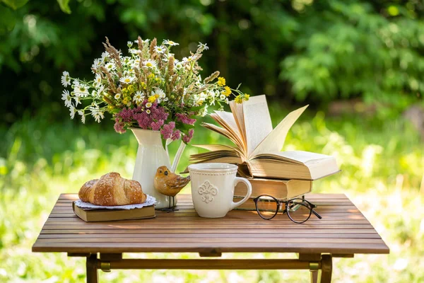 Bouquet of meadow flowers, croissant, cup of tea or coffee, books on table in summer idyllic garden. Rest in garden, reading books, breakfast, vacations in nature concept. Summertime in garden on backyard