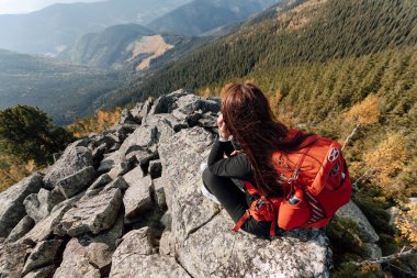 Kırmızı sırt çantalı bir kadın dağların arka planında büyük bir taşın üzerinde oturur. Yalnız seyahat etmek. Kız tepeye tırmandı ve uzaklığa baktı..