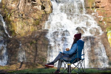 Kadın termostan çay ya da kahve içiyor, şelalenin yanındaki şezlongda oturuyor.. 