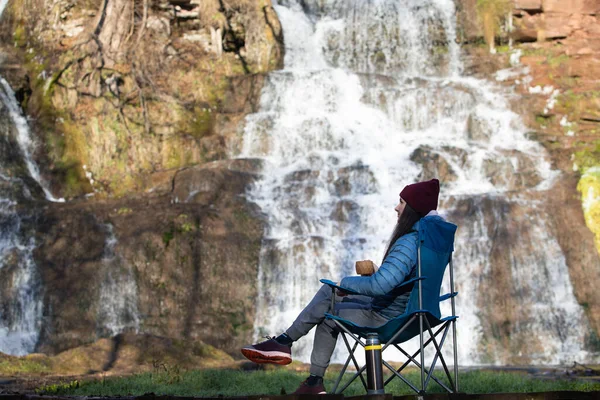 Kadın termostan çay ya da kahve içiyor, şelalenin yanındaki şezlongda oturuyor.. 