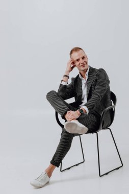 Genç adam bacak bacak üstüne atmış ve beyaz stüdyo arka planında bir takım elbise giyiyor.