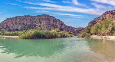 Preveli Girit 'teki nehir geçidinin manzarası turkuaz su ve mavi gökyüzünün altındaki kayalık tepeler