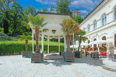 Güneşli bir yaz gününde Karlovy Vary Çek Cumhuriyeti 'nde bir kafe terası yakınında palmiye ağaçlarıyla çevrili beyaz bir spa köşkü..