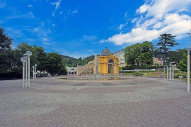 Marianske Lazne 'de çevresi ağaçlar ve tepelerle çevrili, yuvarlak çeşmeli ve açık plazalı ünlü sarı spa salonunun merkezi manzarası..