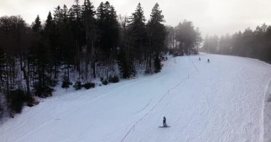 Avusturya Hochficht 'te kayak pistinde kayak yapan kayakçıların orman ve dramatik kış bulutlarıyla çevrili hava manzaralı videosu.