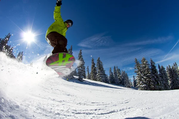 Um homem voando pelo ar enquanto monta um snowboard abaixo de uma colina nevada — Fotografia de Stock