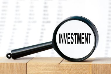 İş adamının elinde INVESTMENT yazılı bir kart tutmasına yakın plan. Yumuşak odaklı iş konsepti görüntüsü. Sayılar sütununun arka planında büyüteç.