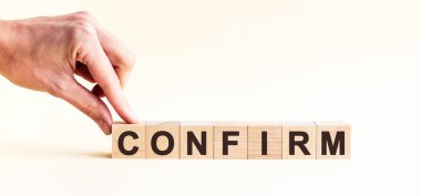 El, CONFIRM kelimesinin C harfinin olduğu tahta bir küp koyuyor. Kelime, masanın sarı yüzeyinde duran tahta küplere yazılmıştır..