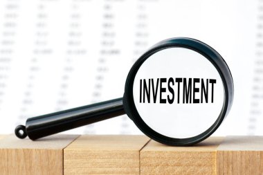 İş adamının elinde INVESTMENT yazılı bir kart tutmasına yakın plan. Yumuşak odaklı iş konsepti görüntüsü. Sayılar sütununun arka planında büyüteç.