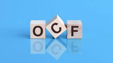 Tahta küpler üzerine OCF metni, hisse senedi konsepti. OCF harfli üç tahta küp. Nakit Akış Operasyonu 'nun kısaltması. Küpler mavi cam bir yüzey üzerinde yer alıyor. OCF - bu şirketin işletim faaliyetlerinden gelen nakit para.