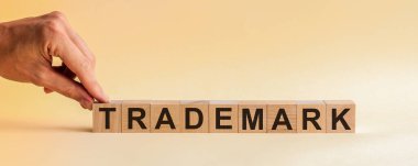 El, üzerinde ticari marka kelimesinin T harfi olan tahta bir küp koyuyor. Kelime, masanın sarı yüzeyinde duran tahta küplere yazılmıştır..