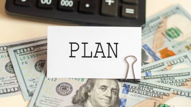 Plan kelimesi beyaz kartta yazılı. Arkaplanda 100 dolarlık banknotlar ve hesap makinesi var.