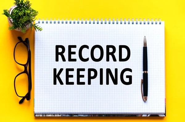 Word text RECORD KEEPING on white paper card, black letters. Pen, glasses and green flower on yellow background. Business concept. Education concept.