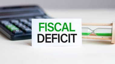 Notlar, hesap makinesi ve beyaz arkaplanda kum saati için FISCAL DEFICIT kağıdı yazılı beyaz kart. İş konsepti. Kahverengi arkaplan.