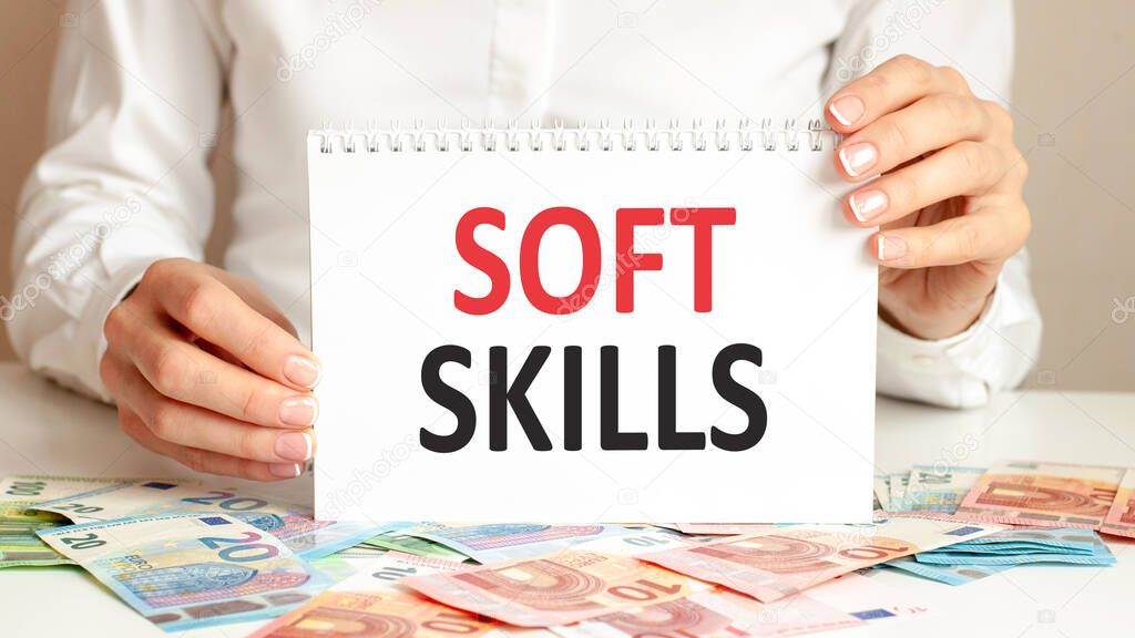 A woman in a white shirt holds a piece of paper with the text: SOFT SKILLS. Business concept for companies and educational institutions. Banknotes and tablet on a table, background.