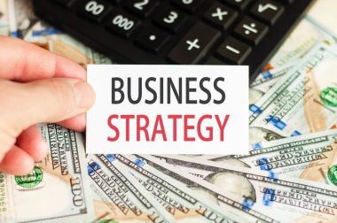 Bir el, banknotların arka planında BUSINESS STRATEGY ve masanın üzerinde bir hesap makinesi yazılı bir tabela tutar. Finans ve ekonomi kavramı.
