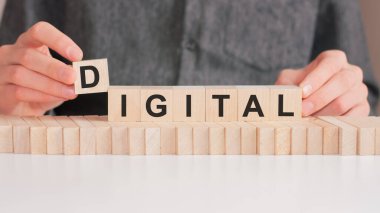 El, Dijital kelimesinin D harfinin olduğu tahta bir küp koyuyor. Kelime, masanın beyaz yüzeyinde duran tahta küplere yazılmıştır..