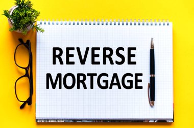 Word text REVERSE MORTGAGE on white paper card, black letters. Pen, glasses and green flower on yellow background. Business concept. Education concept.
