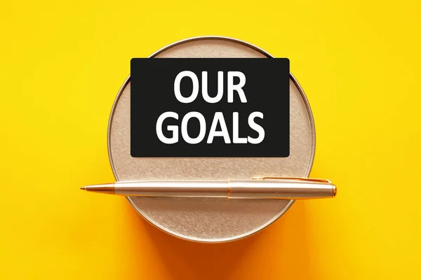 OUR GOALS - words writing a white letters on a sheet paper. Black card with text on a yellow background with round metal stand and metal writing pen. Business, finance and education concept