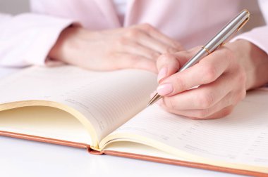 Ahşap masaya beyaz boş bir defter yazan bir kadının yakın plan resmi. Pembe arka plan, seçici odaklanma. İş ve eğitim kavramı.