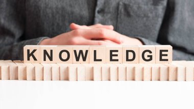 El, KNOWLEDGE harfinin yanına tahta bir küp koyar. Kelime, masanın beyaz yüzeyinde duran tahta küplere yazılmıştır..