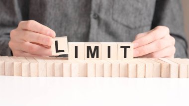 El, LIMIT kelimesinin L harfinin olduğu tahta bir küp koyuyor. Kelime, masanın beyaz yüzeyinde duran tahta küplere yazılmıştır..